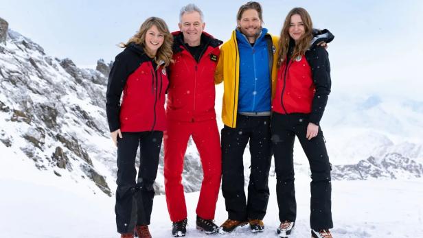 Vier Personen in wetterfester Bergrettungskleidung posieren vor einer schneebedeckten Bergkulisse.