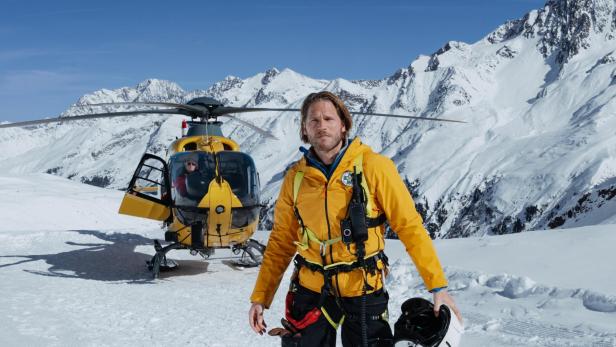 Ein Bergretter in gelber Jacke steht mit einem Helm in der Hand vor einem gelben Hubschrauber in verschneiter Berglandschaft.