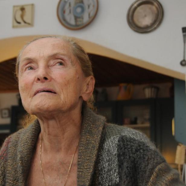 Erni Mangold mit nachdenklichem Blick trägt eine Strickjacke vor einer Wand mit dekorativen Tellern und Bildern.