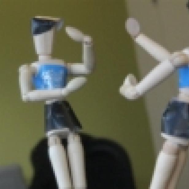 Zwei hölzerne Gliederpuppen, eine sitzend und eine stehend, posieren.