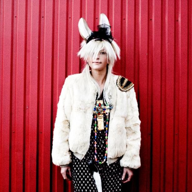 Eine Person mit Hasenohren, Pelzjacke und auffälligem Schmuck vor einer roten Wand.