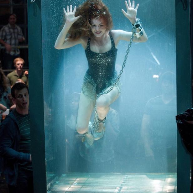Eine Frau, gefesselt in einem Wassertank, wird von Zuschauern beobachtet.