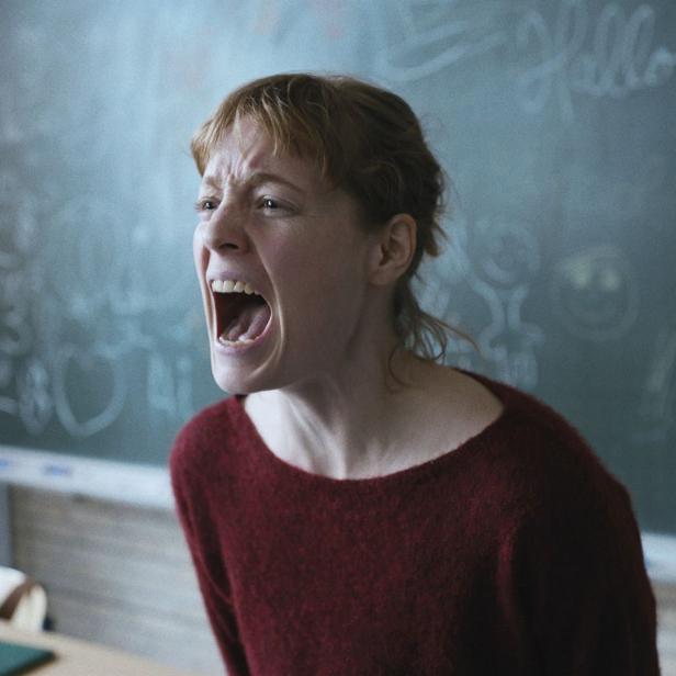 Eine schreiende Frau steht vor einer Tafel in einem Klassenzimmer.