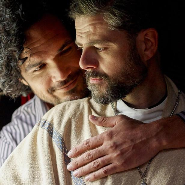 Ein Mann umarmt einen anderen Mann mit Bart vor einem Fenster.