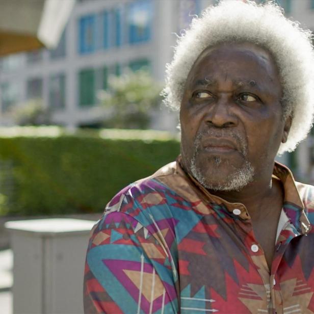 Ein älterer, dunkelhäutiger Mann mit weißem Haar und buntem Hemd blickt nach links.