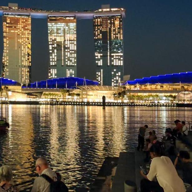 Das Marina Bay Sands Hotel und andere Gebäude spiegeln sich nachts im Wasser, während Menschen am Ufer sitzen.