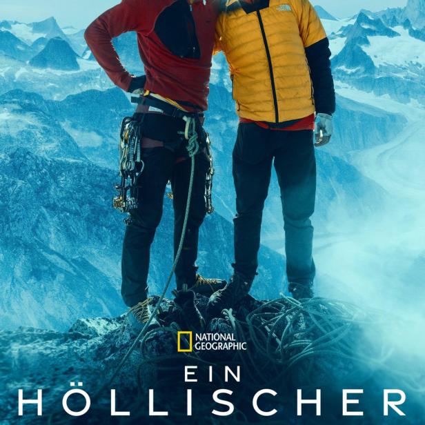 Zwei Bergsteiger stehen auf einem Gipfel mit schneebedeckten Bergen im Hintergrund.