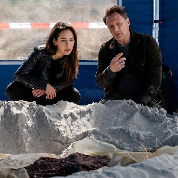 Zwei Personen untersuchen einen Leichnam im Sand, möglicherweise am Tatort eines Nordseekrimis.
