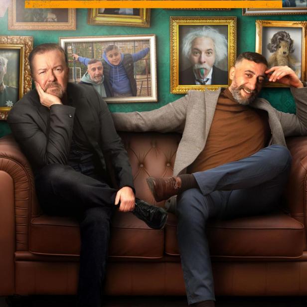 Ricky Gervais und Christoph Maria Herbst sitzen auf einer Couch vor einer Wand mit gerahmten Bildern für „German Genius“.