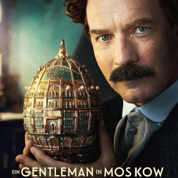 Ewan McGregor hält ein goldenes Miniaturmodell eines Hotels in der Hand.