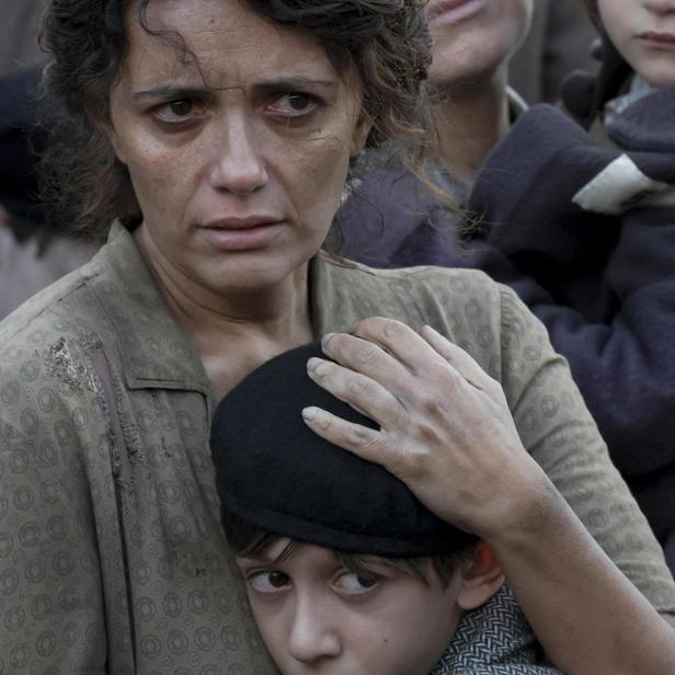 Eine besorgte Mutter umarmt ihren Sohn; im Hintergrund weitere Menschen.