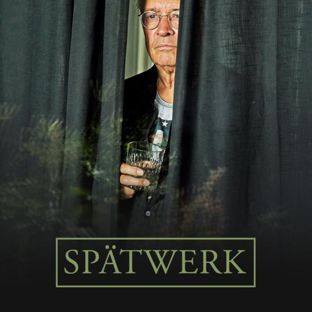 Ein älterer Mann mit Brille hält ein Glas Wasser hinter einem Vorhang.