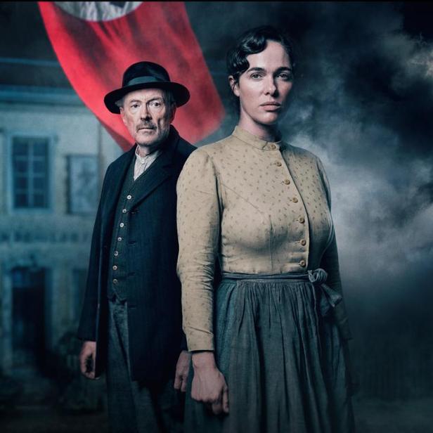 Frau und Mann stehen vor einem Haus unter einer Hakenkreuzfahne mit düsteren Wolken im Hintergrund