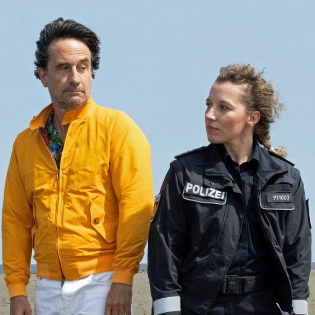 Frau in Polizeijacke und Mann in oranger Jacke blicken einander auf einem Strand an