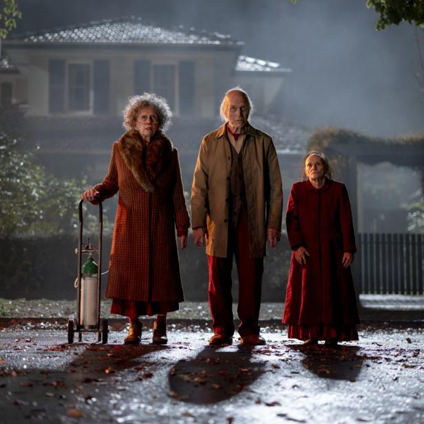 Zwei alte Frauen und ein alter Mann stehen nachts vor einem dunklen Haus auf einer laubbedeckten Straße 