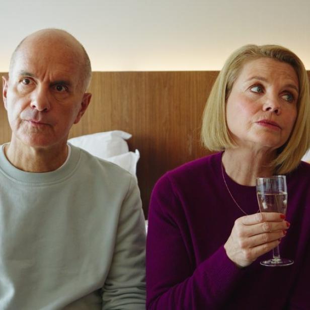 Ein älteres Paar sitzt nebeneinander auf einem Bett, beide schauen ernst, die Frau hält ein Glas mit Getränk.