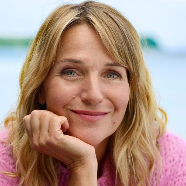 Eine Frau mit blondem Haar und rosa Pullover lächelt am Wasser, daneben ist eine stilisierte Illustration eines Leuchtturms.