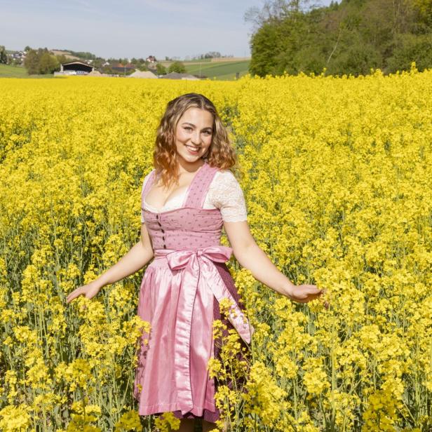 Eine junge Frau im Dirndl steht lächelnd in einem blühenden gelben Rapsfeld bei sonnigem Wetter.