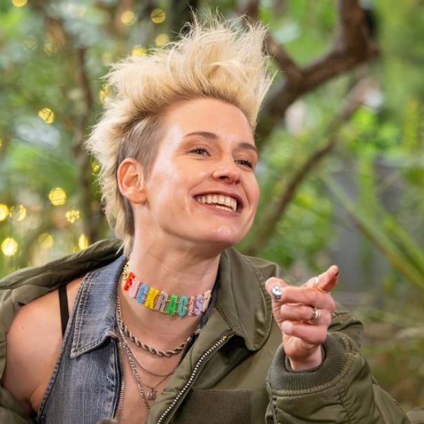 Eine lächelnde Frau mit blondem Irokesenschnitt trägt eine grüne Jacke und eine bunte Halskette.