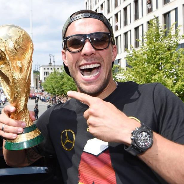 Ein jubelnder Lukas Podolski mit Sonnenbrille hält den WM-Pokal in der Hand und zeigt darauf.