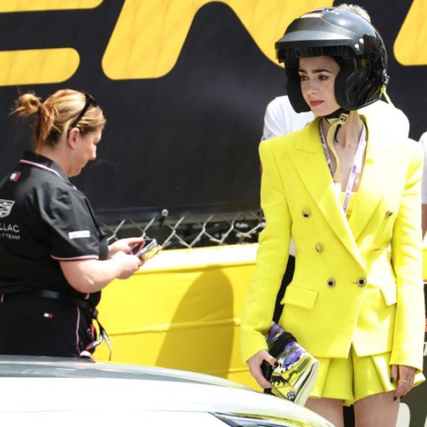 Lily Collins trägt einen gelben Hosenanzug und einen Rennhelm an einer Formel-1-Strecke, während eine Cadillac-Mitarbeiterin daneben steht.
