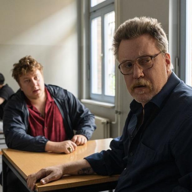 Ein Mann mit Schnurrbart und Brille sitzt in einem Raum mit vergitterten Fenstern, während im Hintergrund zwei weitere Personen an einem Tisch sitzen.