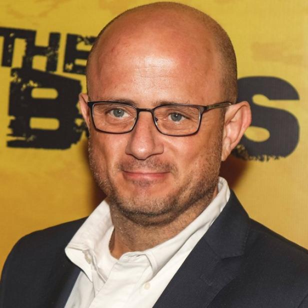 Ein Mann mit Brille und Glatze trägt ein dunkles Sakko und steht vor einem gelben Hintergrund mit schwarzer Schrift.