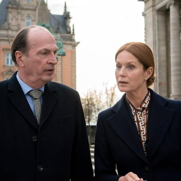 Ein Mann und eine Frau in dunklen Mänteln unterhalten sich vor einem historischen Gebäude.