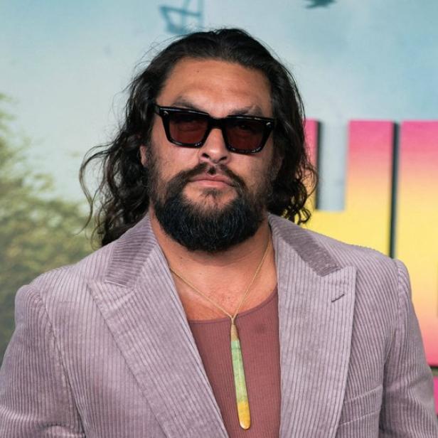 Jason Momoa trägt eine Sonnenbrille, ein mauvefarbenes Cord-Sakko und eine lange Halskette vor einem bunten Hintergrund.