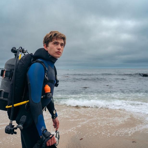 Ein Taucher in einem blauen Neoprenanzug mit Ausrüstung steht an einem bewölkten Tag am Strand vor dem Meer