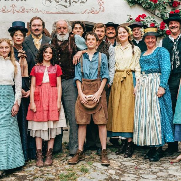 Eine große Gruppe von Menschen in historischer Kleidung posiert vor einem Haus mit einer Rosenranke an der Wand.