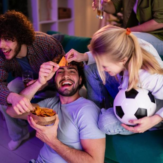 Eine Gruppe junger Freunde sitzt auf einem Sofa, isst Chips und schaut gemeinsam Fußball.
