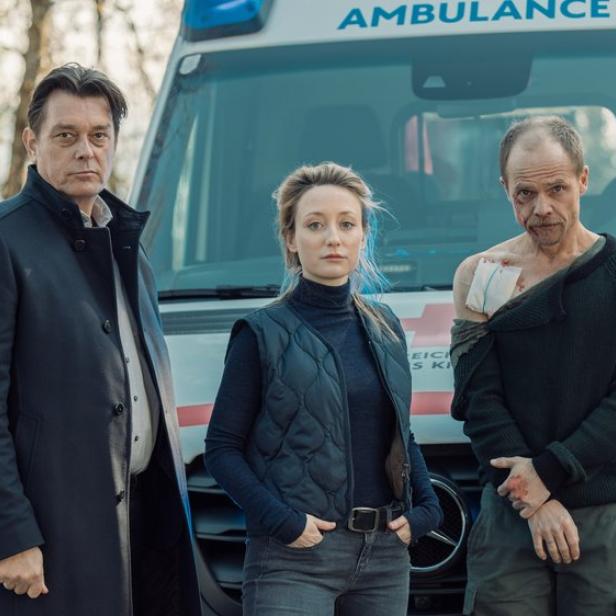 Drei Personen stehen vor einem Krankenwagen, eine davon hat eine sichtbare Verletzung an der Schulter.