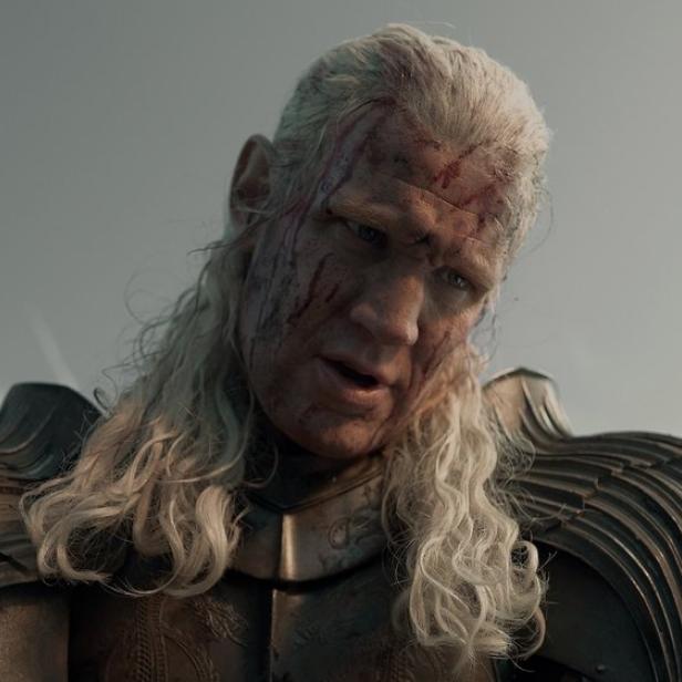 Matt Smith als Daemon Targaryen mit blutverschmiertem Gesicht und langem, hellem Haar in einer Rüstung vor einem grauen Himmel.