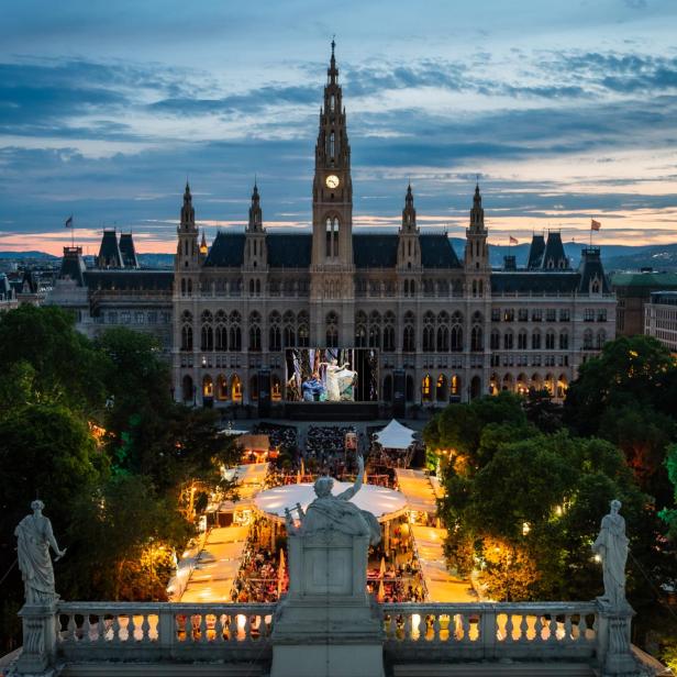Das Wiener Rathaus bei Abenddämmerung mit einer großen Leinwand und beleuchteten Ständen auf dem Rathausplatz.
