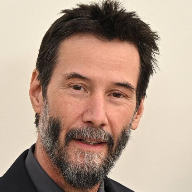 Keanu Reeves mit dunklem Haar und Bart vor einem hellen Hintergrund.