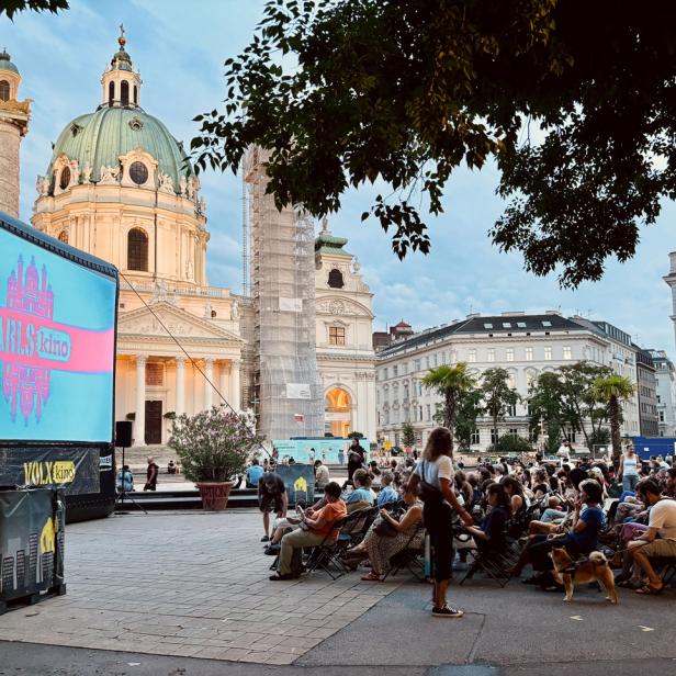 Zuschauer sitzen vor einer großen Leinwand des Volxkino auf dem Wiener Karlsplatz mit der Karlskirche im Hintergrund.