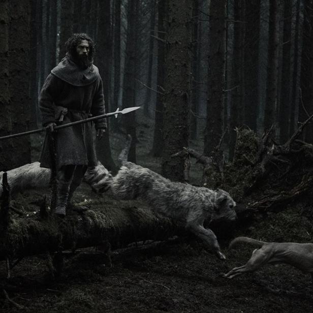 Ein bärtiger Mann in mittelalterlicher Kleidung mit einem Speer steht in einem dunklen Wald, während zwei Hunde über einen umgestürzten Baum springen.