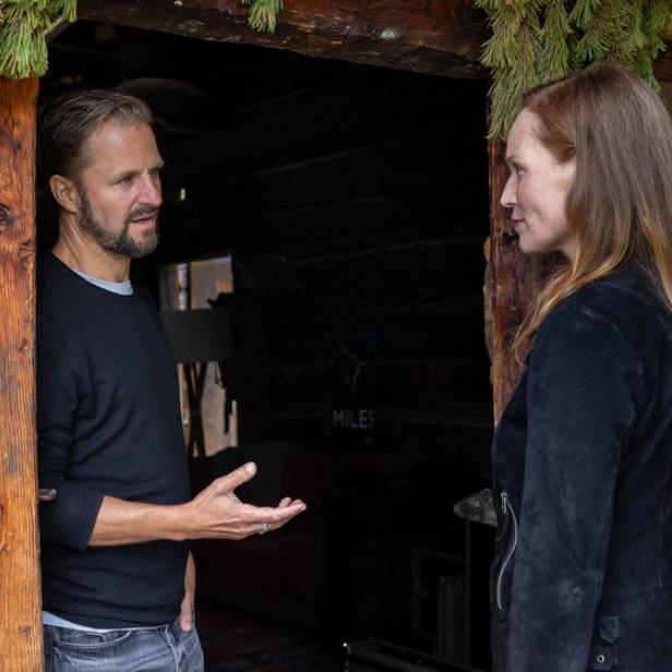 Ein Mann und eine Frau unterhalten sich vor dem Eingang einer rustikalen Holzhütte.