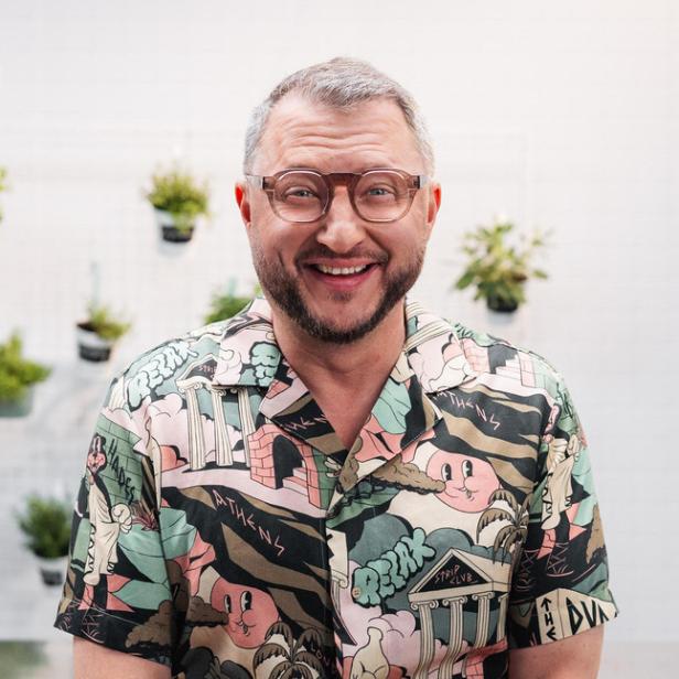 Ein lächelnder Mann mit Brille trägt ein buntes Hemd mit Comic-Motiven vor einer Wand mit hängenden Pflanzen.