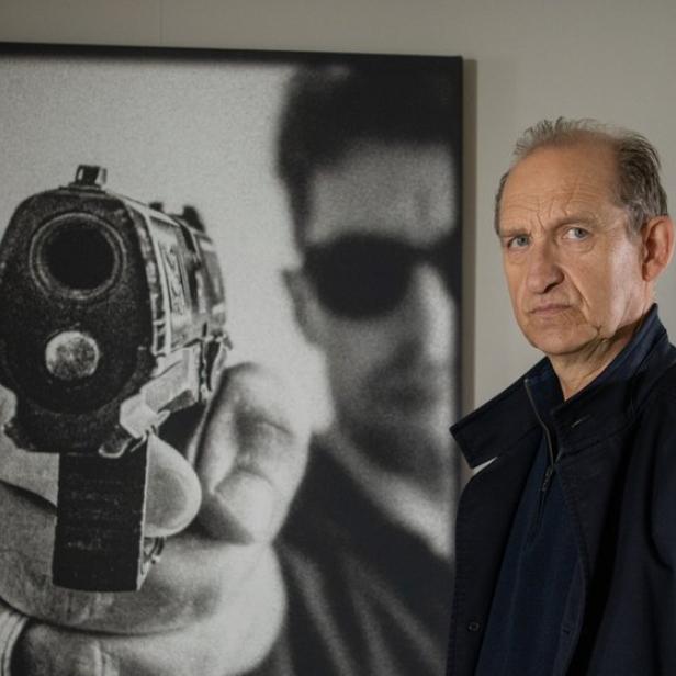 Ein älterer Mann mit ernstem Blick steht vor einem schwarz-weißen Wandbild, das eine Person mit einer auf den Betrachter gerichteten Pistole zeigt.