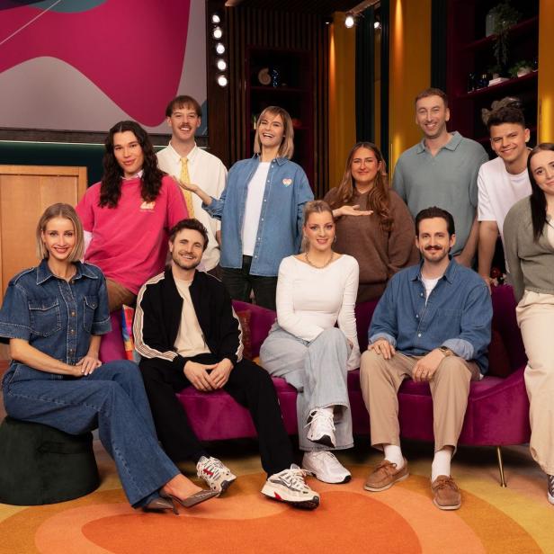 Eine Gruppe von elf jungen Erwachsenen posiert gemeinsam in einem farbenfrohen Fernsehstudio vor einem violetten Sofa.