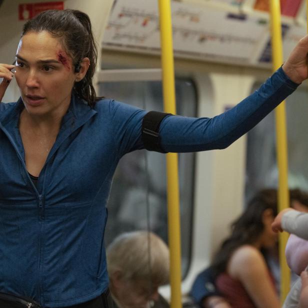 Gal Gadot mit einer Kopfverletzung telefoniert in einer U-Bahn, während sie sich an einer gelben Stange festhält.