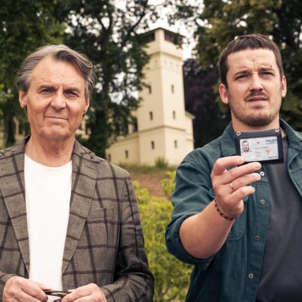 Zwei Männer stehen vor einem Turm, wobei einer von ihnen einen Polizeiausweis in die Kamera hält.