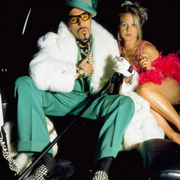 Ein Mann in grünem Anzug mit weißem Pelzmantel und eine Frau in einem pinken Kleid sitzen mit einer Champagnerflasche in einer Limousine.