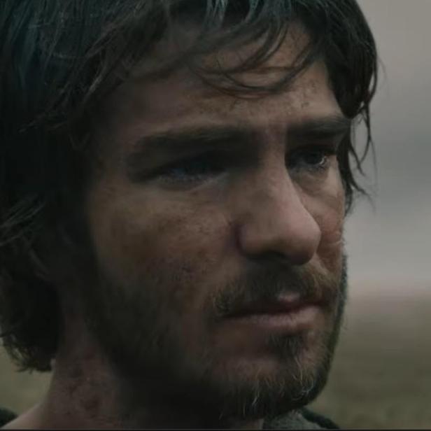 Andrew Garfield blickt mit ernstem, schmutzigem Gesichtsausdruck in einer trüben, weiten Landschaft in die Ferne.