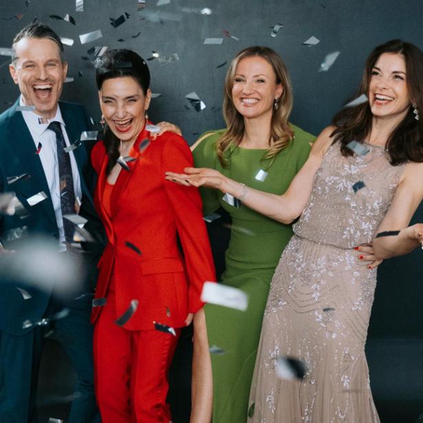 Vier festlich gekleidete Personen lachen vor einem dunklen Hintergrund, während silbernes Konfetti auf sie herabfällt.