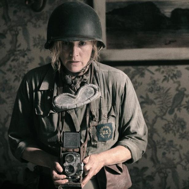 Kate Winslet trägt als Kriegskorrespondentin Lee Miller einen Helm und hält eine Kamera vor dem Hintergrund einer gemusterten Tapete.
