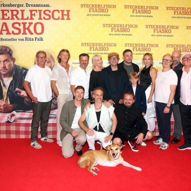 Gruppenfoto auf dem roten Teppich vor dem Filmplakat „Steckerlfisch Fiasko“, mit mehreren Prominenten und einem liegenden Hund.