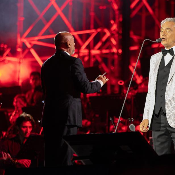 Andrea Bocelli singt vor einem Orchester, während ein Dirigent den Auftritt vor einer rot beleuchteten Bühne leitet.